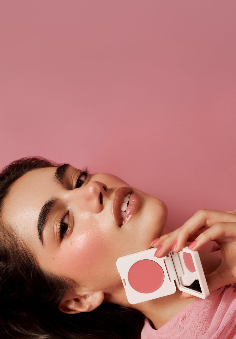Woman applying blush to her cheeks with a pink background