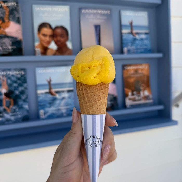 Hand holding a yellow ice cream cone with a branded container against a blue wall with framed pictures.