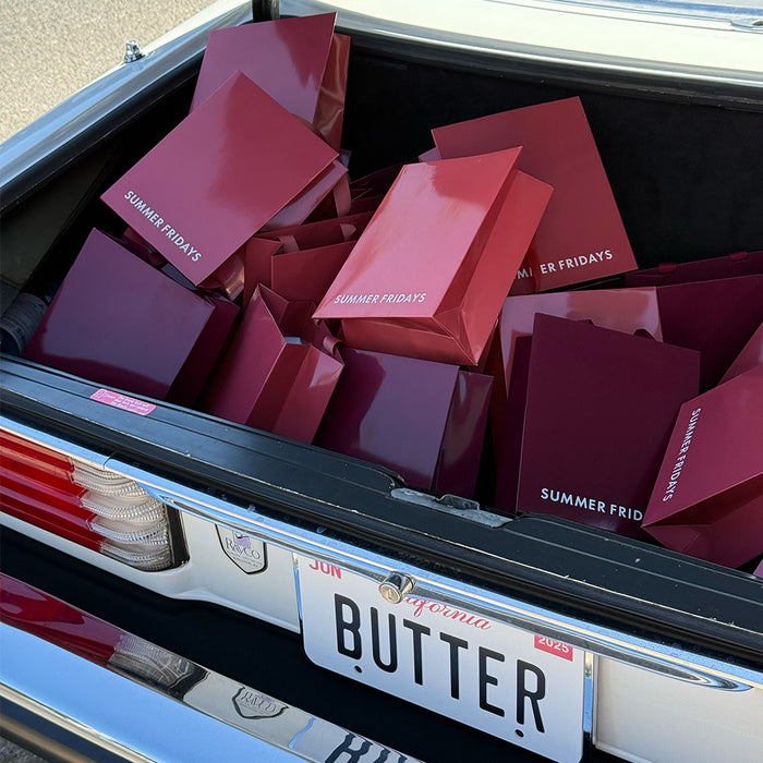 Red 'Summer Fridays' shopping bags in a car trunk with a license plate reading 'BUTTER'.