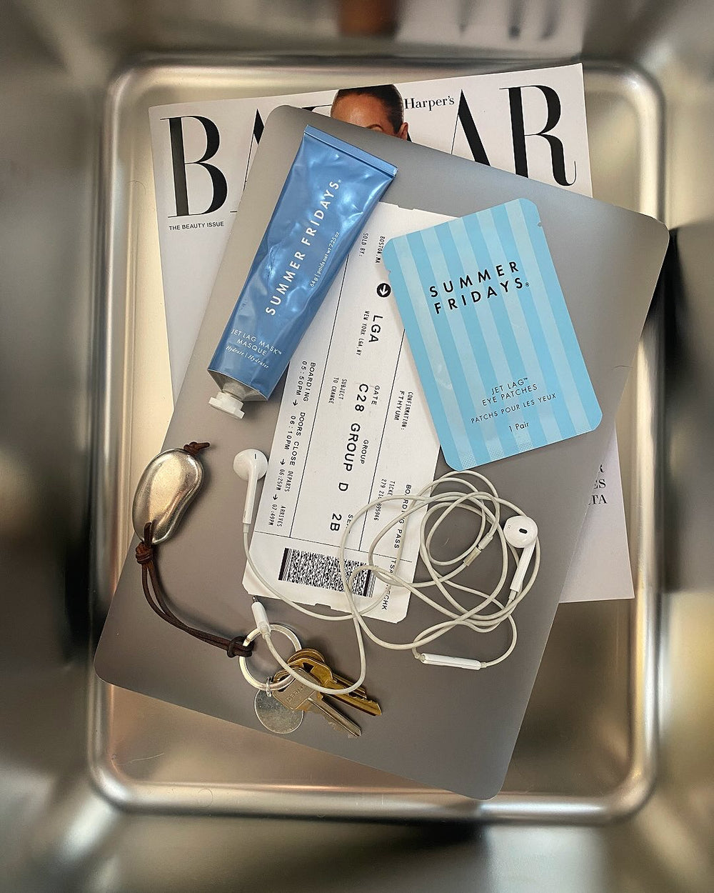 Different items including a Jet Lag Mask, Jet Lag Eye Patches, a plane ticket, headphones, keys and a computer in side a silver bin.