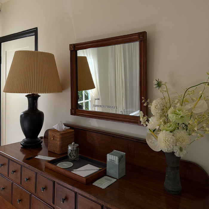 Wooden dresser with mirror, lamp, tissues, and flowers in a room.