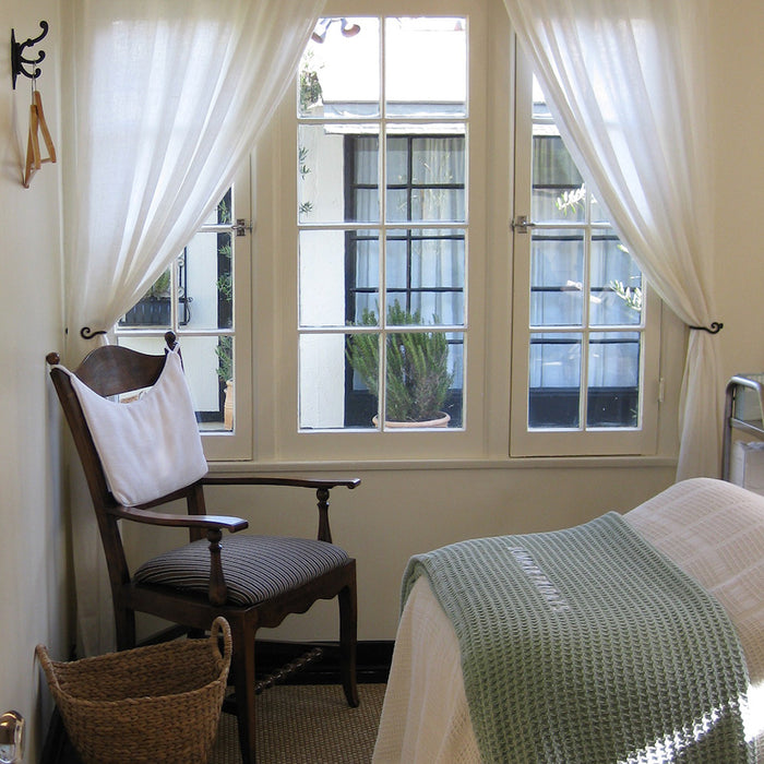 Room interior with a chair, basket, and bed with a green blanket near a window with white curtains.