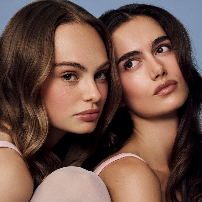 Two women with long hair wearing pink tank tops against a blue background