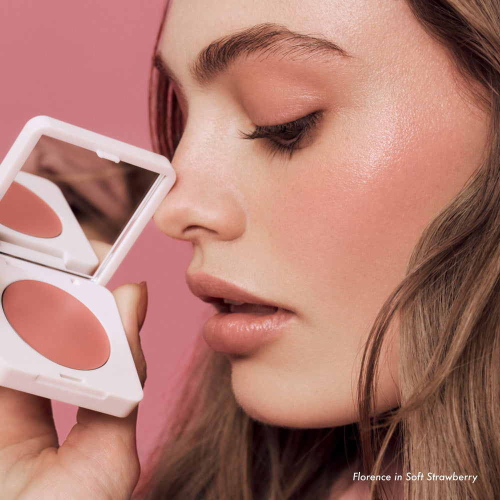Close-up of a woman applying blush with a compact mirror against a pink background