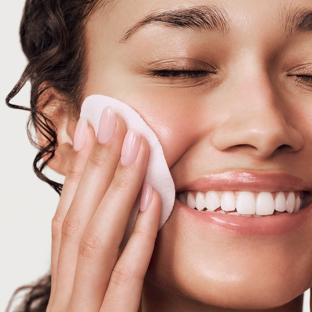 Woman using a white facial exfoliating pad on her face against a neutral background