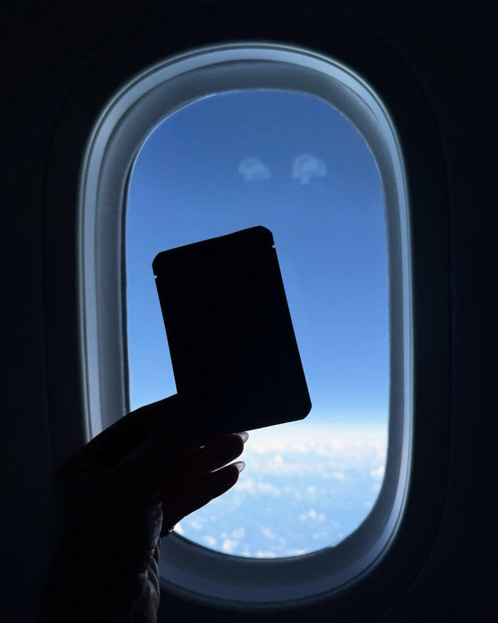 Hand holding a pouch of Jet Lag Eye Patches over a plane window and blue sky