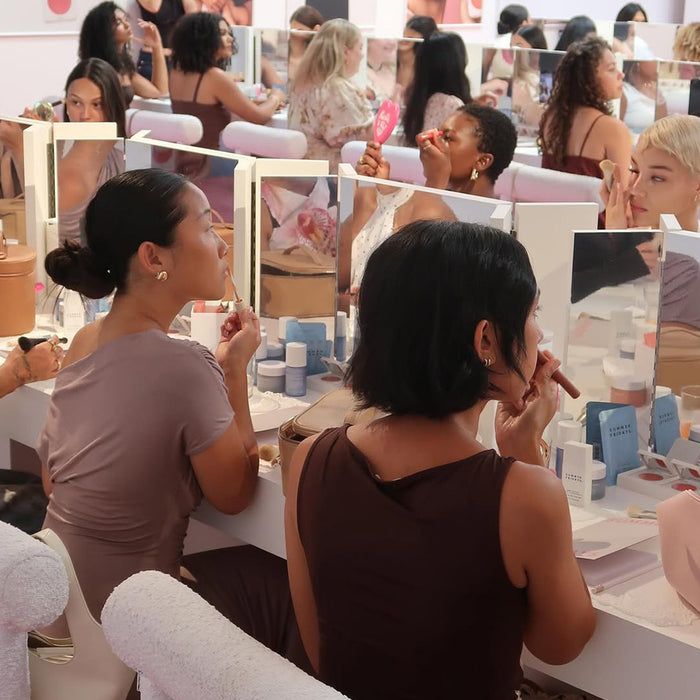 Two women sitting at a table with makeup products and mirrors, surrounded by other people.