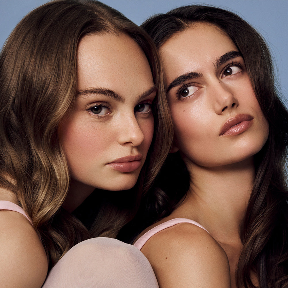 Two women with long hair wearing pink tank tops against a blue background